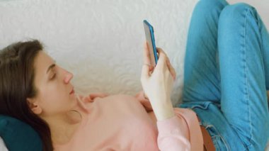 Esmer bir kadın evinde boş bir anın tadını çıkarıyor. Kanepede oturuyor ve akıllı telefonuna tatmin olmuş bir ifadeyle bakıyor..