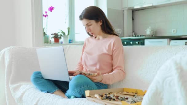 一个年轻的女人一边在舒适的客厅里享受着一片披萨 一边在笔记本电脑上辛辛苦苦地工作 在家里工作的美丽一个年轻的女人带着笔记本电脑舒服地坐在沙发上 — 图库视频影像