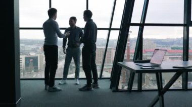 Üç genç ofis çalışanı iş hakkında konuşuyor, bir grup erkek meslektaşı laptoplar üzerinde verimli bir toplantı yapıyorlar.