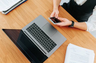 Dizüstü bilgisayarda daktilo yazan ve ofisteki bir masada akıllı telefon tutan adamların ellerini yakın çekim. Üst görünüm, aygıtlar arasında birden fazla görev yapan meşgul ofis çalışanlarını yakalar.