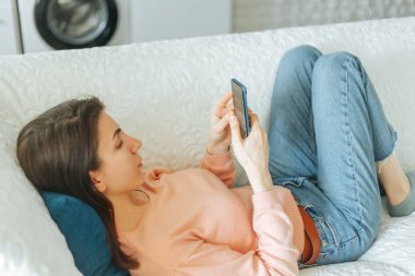 Rahatlamış ve mutlu bir genç kadın, oturma odasında kanepede oturuyor ve sosyal medya hesaplarını akıllı telefonuyla karıştırıyor..