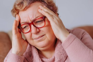 Elderly woman struggling with a severe headache, her hands on her head, while sitting in her comfortable living room, Dealing with Difficulty Elderly Woman Battles Aches and Pains