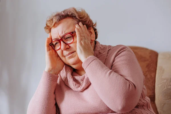 Kıdemli bir kadın, elleri başının üstünde, baş ağrısıyla boğuşuyor, evde zonkluyor, baş ağrılarıyla boğuşuyor, kıdemli kadın evde destek buluyor.