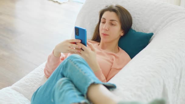 自宅のリビングルームでソファに座りながらスマートフォンの最新ファッショントレンドをオンラインで購入する若い女性 — ストック動画