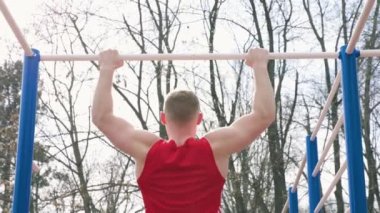 Terleme ve Dayanıklılık Etkinliği olan adam açık hava spor salonunda barfiks çekiyor. Aktif bir adam spor salonunun barında barfiks çekiyor. Kırmızı tişörtü terden sırılsıklam olmuş..