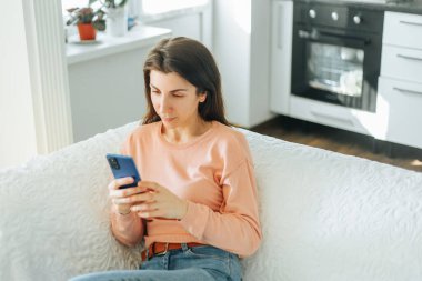 Çekici genç bir kadın bu fotoğrafta görülüyor, kanepesinde oturuyor ve akıllı telefonuyla sosyal medyada geziniyor, mutlu ve mutlu görünüyor..