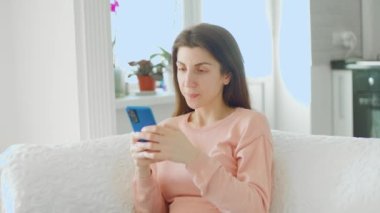 Çekici genç bir kadın bu fotoğrafta görülüyor, kanepesinde oturuyor ve akıllı telefonuyla sosyal medyada geziniyor, mutlu ve mutlu görünüyor..