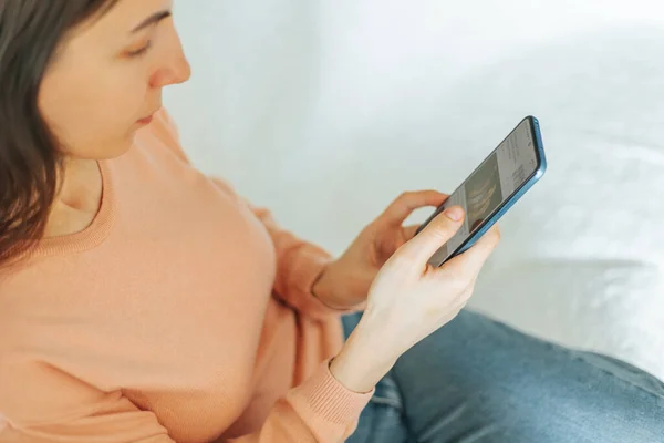 stock image top view, authentic image shows a young woman browsing her smartphone while sitting on her sofa at home, looking relaxed and satisfied.