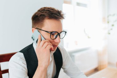 30 'lu yaşlarının başında bir adam, ofisinde oturmuş, akıllı telefonuyla patronuyla konuşuyor. Uzaktan kumandalı bir işçi ve çalışma alanı bir dizüstü bilgisayar, dosyalar ve beyaz tahtayla dolu.