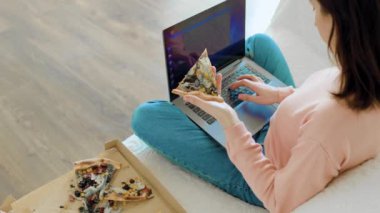 Lezzetli bir dilim pizzanın tadını çıkarmak için ara verdiğinde bir kadın dizüstü bilgisayarında yazı yazıyor. Dizüstü bilgisayarından yeni bir şey öğrenirken bir kadının elinde bir dilim pizza tutarken yakın plan fotoğrafı..