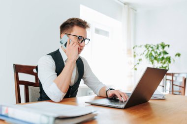 Ofiste, genç bir adam masasının yanında duruyor, akıllı telefonuyla acil bir ses tonuyla konuşuyor. Dizüstü bilgisayarı ve iş belgeleri yakınlarda, beyaz yakalı bir işçi olarak kendini adadığını gösteriyor.