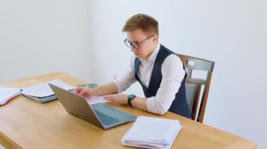 Bir ofis çalışanı iş yerinde oturuyor, etrafı iş arkadaşlarıyla çevrili. Gözlük ve beyaz yakalı gömlek giyerken bir kağıda önemli bilgiler yazıyor..