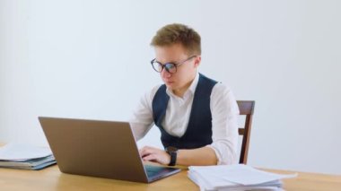 Beyaz yakalı gömlek ve kravat giymiş şık genç bir işadamı, modern ev ofisinde oturur, gözlükleriyle dizüstü bilgisayarına bakar ve not alır..