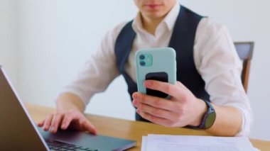 Gözlüklü genç bir beyaz adam akıllı telefonuyla ciddi bir konuşma yapıyor. Genç bir yetişkin ofiste gündelik bir konuşma yapıyor. Akıllı telefonunu bir elinde tutuyor.