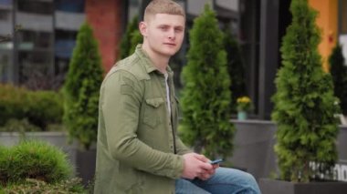 Genç yetişkin erkek beklerken cep telefonuyla kameraya bakıyor. Akıllı telefonuyla poz veren yakışıklı adam. Memnun çocuk şehirde akıllı telefonuyla rahatlıyor.
