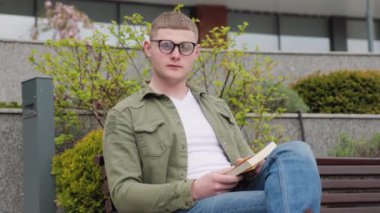 Edebi Urbanite A Young, gözlüklü adam şehirde kitapla oturuyor. Elinde kitap olan genç bir erkek şehir ortamında oturuyor. Portresinde yakışıklı ve otantik görünüyor..