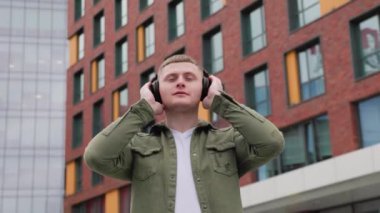 Kulaklıklı yakışıklı bir adam, arka plandaki güzel şehir binalarının manzarasını seyrediyor ve eğleniyor. Şehir Kafiyeli Yakışıklı Adam Kulaklıkla Rahatlıyor