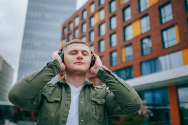Genç bir adamın yüzü, kulaklıkla müzik dinlerken ve şehir manzarasını izlerken gülümserken. Şehir Titreşimleri Yakışıklı Adam Açık havada Müzik Dinliyor
