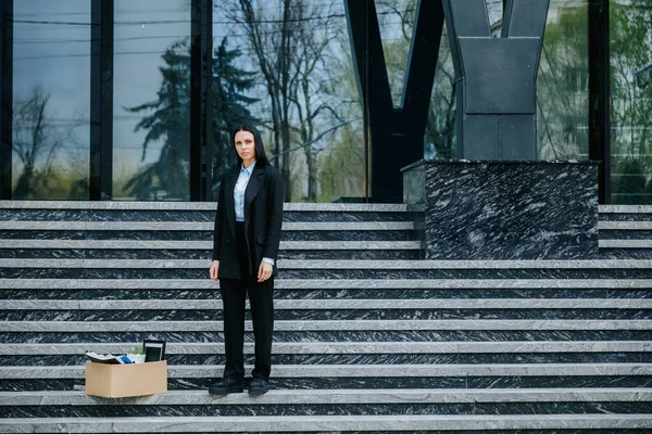 A woman stands on the stairs of a high-rise building with a carton box, reflecting the reality of job loss and unemployment during a time of crisis and economic downturn.