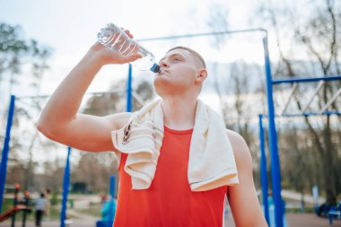 Ter dökerek erkek sporcu dışarıda idman yaparken su içer. Yorgun bir adam su içmek ve temiz havada dinlenmek için spor faaliyetlerine ara verir..