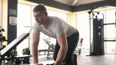 Spor salonunda, genç beyaz bir adam ağırlık kaldırarak spora bağlılığını gösteriyor. Aktif sporcu, fiziksel kondisyonunu geliştirmek için yüksek yoğunluklu kapalı alanda antrenman yapıyor.