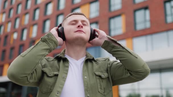 一个拿着耳机的年轻人 抬头看着城市的天际 一边听着他最喜欢的音乐 一边笑着 戴耳机的帅小伙松懈 — 图库视频影像