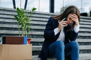 Dışarıda oturan bir kadın, elinde karton levhayla akıllı telefonuyla konuşuyor, kayıp, işsizlik deneyimlerini ve iş bulma zorluklarını paylaşıyor..