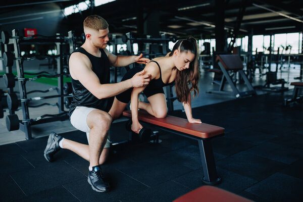 In the gym, a skilled male personal trainer aids a woman in her weightlifting journey, specifically focusing on her dumbbell lift and technique.