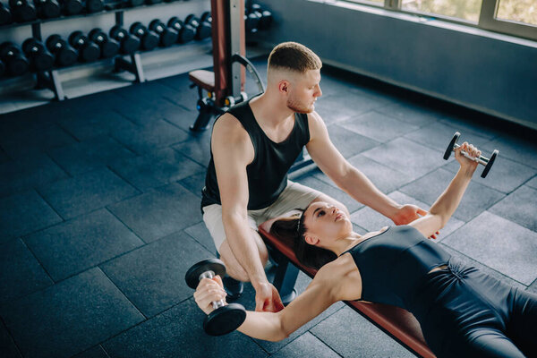 In the gym, a knowledgeable male personal trainer provides guidance to a woman lifting a dumbbell. Fitness Training Male Personal Trainer Helps Woman with Dumbbell Exercise