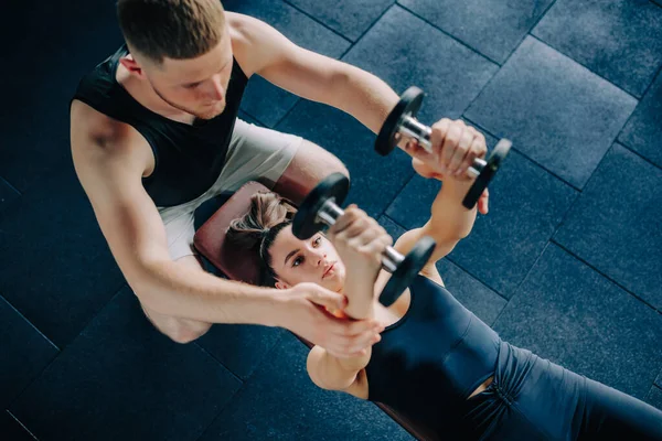 With the support of a male personal trainer, a woman confidently lifts a dumbbell during a gym training session. Dumbbell Exercise Male Personal Trainer Supports Womans Workout