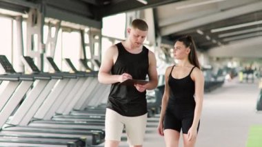 Bir erkek kişisel eğitmen ile bir kadın arasındaki etkileşim, dinamik bir diyalog kurarak, egzersizleri tartışırken ve fitness ile ilgili soruları ele alırken, bu görüntünün merkezinde yer alıyor..