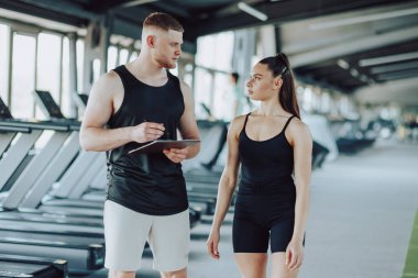 Bir erkek kişisel eğitmen ile bir kadın arasındaki etkileşim, dinamik bir diyalog kurarak, egzersizleri tartışırken ve fitness ile ilgili soruları ele alırken, bu görüntünün merkezinde yer alıyor..