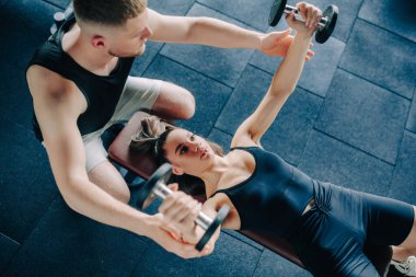 Spor salonunda, destekleyici bir erkek antrenör bir kadına bir dambılı kaldırırken rehberlik eder. Atletik Destek Erkek Kişisel Eğitmen Halter Kadın YardımcısıName