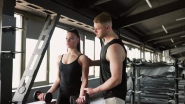 Erkek bir özel antrenör, bir kadının bir dambılı kaldırmasına yardım eder, spor salonunda kişisel eğitim sağlar. Fitness Partnerliği Erkek Kişisel Eğitmeni Aptal Kadın Kalkmasına Yardım Ediyor