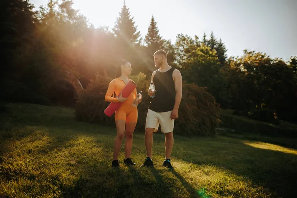 stock image Two athletes, dressed in fitness clothes, are standing outdoors, talking and relaxing after training. The man is holding a bottle of water, and the woman is holding a fitness mat.