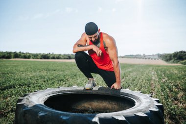 Formda ve kararlı bir yüz ifadesi olan bir adam antrenmandan sonra Cross Fit tekerleğinin yanında mola verir. Çapraz Fit Sporcusu Yoğun Eğitimden Sonra Dışarıda Rahatlıyor