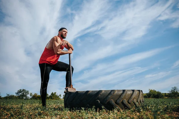 Strong Male Athlete with the hammer After Cross Fit Training Outdoors. Portrait of a strong Caucasian man resting by the CrossFit wheel after a challenging workout outdoors.