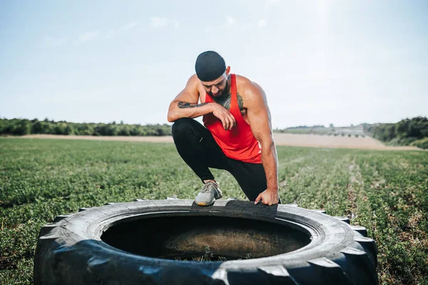 Fit man with a determined expression takes a break by the Cross Fit wheel after training outside. CrossFit Athlete Relaxing Outdoors After Intense Training