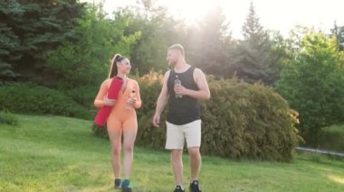İki sporcu, spor kıyafetleri giymiş genç bir erkek ve bir kadın, antrenmandan sonra dışarıda durup konuşuyorlar ve dinleniyorlar. Adam bir şişe su tutuyor, kadın da bir spor paspası tutuyor..