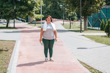 Büyük beden bir kadın gülümsemesi, açık hava eğitiminden sonra onun mutluluğunu ve memnuniyetini yansıtır. Elinde bir su şişesi ve havlu tutarak hak ettiği molayı alıyor..