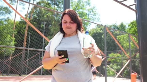 一个身材超大的女人 在完成了户外训练后 在使用智能手机时找到了快乐 她满意地笑着 拿着水瓶喝水 — 图库视频影像