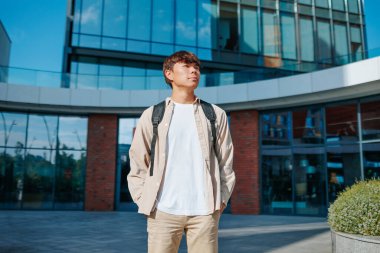 Üniversite bahçesinin huzurlu vahasında, Asyalı bir genç, okul çantası hazır, bilgi dünyasına dalmak, doğayı ve eğitimi harmanlamak için mükemmel bir ortam buluyor..