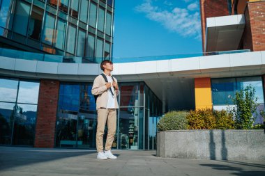 Şehir arkaplanı, yanında okul çantası olan odaklanmış Asyalı bir üniversite öğrencisi, üniversite bahçesinin doğal güzelliğinin tadını çıkarırken, çalışmalarının karmaşıklığı üzerine kafa yoruyor.