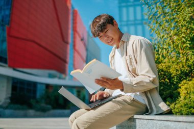 Teknoloji ve Gelenek doğanın kucağında, teknoloji meraklısı genç bir adam dijital ve geleneksel öğrenme arasında uyumlu bir denge sağlar. Dizüstü bilgisayar açık, kitap elinde, tutkuyla çalışıyor..