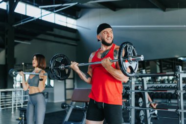 Atletizm ruhuyla dolu bir spor salonunda hem genç hem de Kafkas kökenli bir erkek ve kadın fitness hedeflerine ulaşmak için ağırlık kaldırıyor..