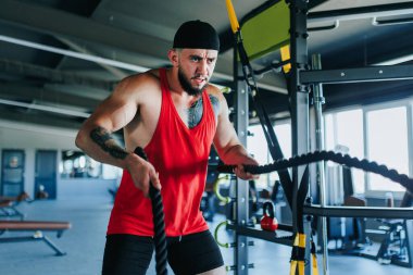 Konsantre ve kararlı bir genç adam halat egzersizini kapalı spor salonunda yapıyor. Kaslı fiziği, kendini adamışlığı ve atletizmiyle birleşmiş.,