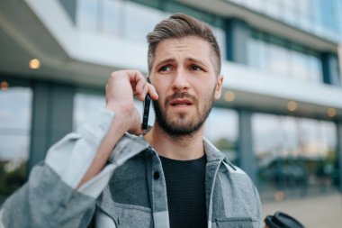 Genç sakallı bir adam şehirde bir telefon görüşmesi yaparken üzgün görünüyor. Cep telefonlu kızgın adam, Konuşmanın derinliklerinde, üzgün görünüyor.
