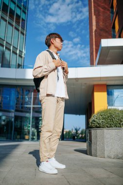 Dikey çekim. Huzurlu bir portre, sakin bir üniversite bahçesinde açık hava öğrenme fırsatını yakaladığı için okul çantası harekete hazır genç bir Koreli öğrenciyi gözler önüne seriyor..