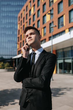 Açık bir şehir ortamında, beyaz yakalı genç bir erkek, iş elbisesi giymiş, akıllı telefonuyla konuşuyor, genç beyaz yakalı bir işçinin günlük yaşamını vurguluyor..