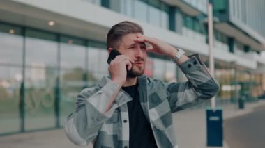 Açık hava ortamında, genç sakallı bir adam ciddi ciddi akıllı telefonuyla konuşuyor. Hoşnutsuz Genç Adam Akıllı Telefonunda Sorunları Tartışıyor